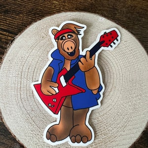 May include: A cartoon character with brown fur, a red headband, and a blue shirt is playing a red electric guitar. The character has a large, brown nose and a wide smile.