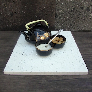 May include: A white square serving tray with black speckled flecks. A black teapot with a gold design and a light green handle sits on the tray. Two small black bowls with white rims are on the tray, one filled with white sugar and the other with small brown cookies. A silver spoon rests on the tray.