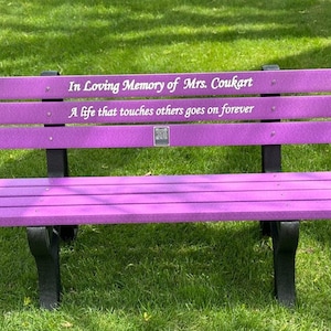 May include: Purple wooden bench with black metal legs. The bench has a plaque with the inscription "In Loving Memory of Mrs. Coukart. A life that touches others goes on forever."