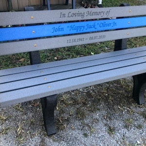 May include: A gray wooden bench with a blue stripe and the inscription "In Loving Memory of John "Happy Jack" Oliver Jr. 12.18.1941 - 08.07.2020"