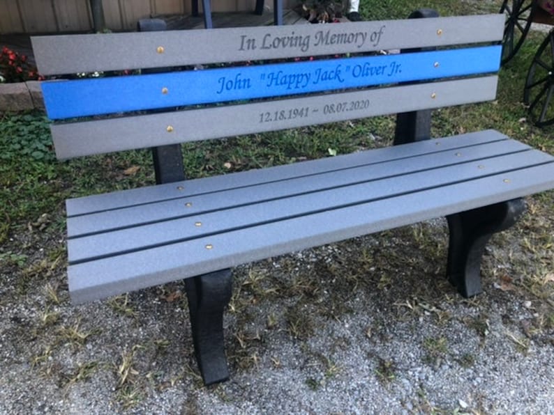 May include: A gray wooden bench with a blue stripe and the inscription "In Loving Memory of John "Happy Jack" Oliver Jr. 12.18.1941 - 08.07.2020"