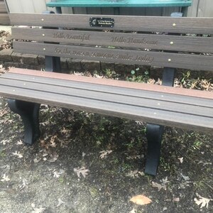 May include: A brown wooden bench with black metal legs. The bench has a plaque on the back with the text "Hello Beautiful, Hello Handsome, Be the reason someone smiles today."