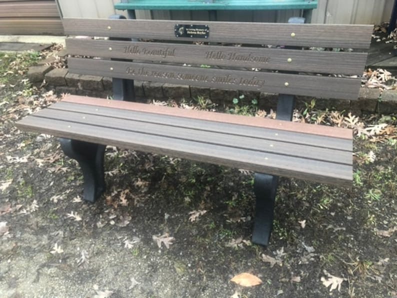 May include: A brown wooden bench with black metal legs. The bench has a plaque on the back with the text "Hello Beautiful, Hello Handsome, Be the reason someone smiles today."