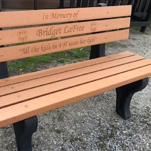 May include: A memorial bench with a tan seat and backrest featuring black legs. The backrest is engraved with the text In Memory of Bridget LaFree, 4-H logos, and the phrase All the kids in 4-H were her kids. The bench is constructed with horizontal slats.