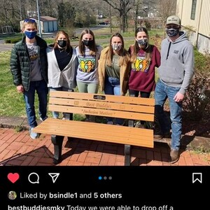 May include: A group of people stand in front of a wooden bench with the words "Best Buddies" and "Inclusion Matters" engraved on it. The bench is a memorial for a late friend.