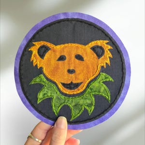 Könnte beinhalten: Ein runder, schwarzer Stoff-Patch mit einem violetten Rand. Der Patch zeigt einen lächelnden, orangefarbenen Bären mit einem grünen, blattförmigen Kragen. Der Bär hat schwarze Augen und eine schwarze Nase.
