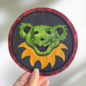 May include: A green and yellow embroidered patch of a smiling bear with a yellow collar. The patch is circular and has a red border.