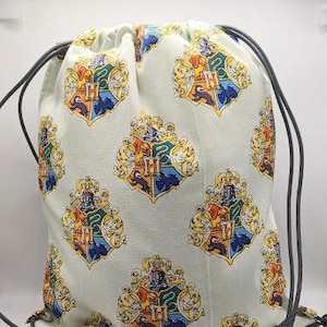 Puede incluir: Una bolsa de cordón blanco con un cordón gris. La bolsa está decorada con escudos de armas coloridos que representan un león, un águila, un tejón y una serpiente.