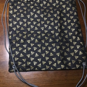 Puede incluir: Mochila negra con cordón y un patrón repetido de triángulos dorados. La bolsa tiene cordones grises y está hecha de tela. El patrón es un símbolo repetido.