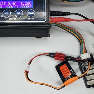May include: A black and red battery charger with a white digital display showing "Li25 0.4A 8.20V RAI AAA:21 00002". The charger is connected to a black and orange battery pack with a white connector. The battery pack is labeled "XH ADAP".