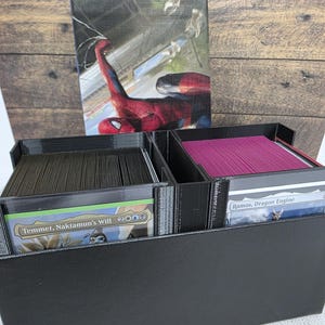 May include: A black plastic trading card storage box with two compartments holding stacks of cards. The left stack shows a card labelled Temmet, Naktamun's Will, and the right stack shows a card labelled Ramos, Dragon Engine. A Spider-Man themed card box lid stands upright behind the storage box.