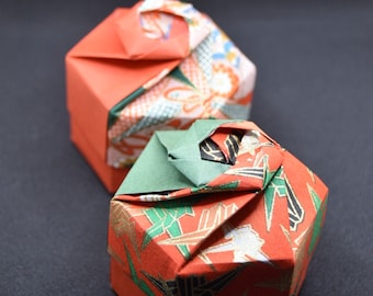 May include: Two origami boxes in the image. The boxes are red with patterned accents in green, gold, and white. The lids are folded in a unique design. The background is a solid black.