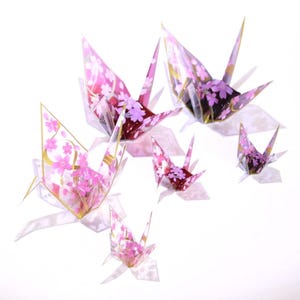 Op de afbeelding: Een groep van zes roze en witte origami-kranen met kersenbloesem-ontwerpen. De kranen zijn gemaakt van papier en hangen aan het plafond.