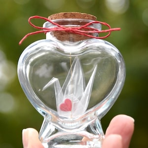 Peut inclure: Une bouteille en verre transparente en forme de cœur avec un bouchon en liège noué avec de la ficelle rouge. À l'intérieur, une grue en origami blanche avec un cœur rouge est suspendue.