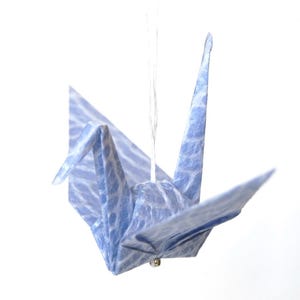 May include: A blue origami crane suspended by a thin white string. The paper crane has a textured, water-like pattern. The crane's wings are spread, and its beak is pointed upwards. A small, gold-colored bead is attached to the bottom.