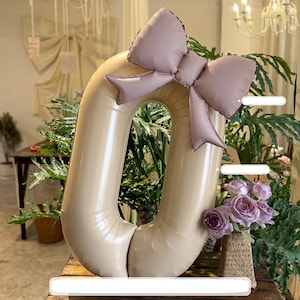 Może przedstawiać: Duży, beżowy balon w kształcie cyfry zero jest ozdobiony malinową kokardą. Balon umieszczono na drewnianej powierzchni, z zielenią i kwiatami w tle. Kokarda wykonana jest z podobnego materiału co balon.