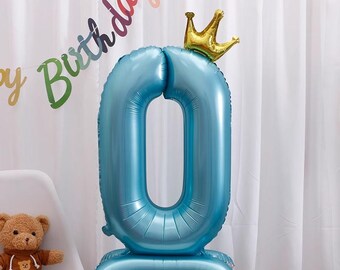 Pode incluir: Um grande balão azul claro em forma do número zero, encimado por uma coroa dourada. O balão está sobre uma base. Uma faixa com a frase "Happy Birthday" é visível ao fundo. Um ursinho de pelúcia está sentado perto.