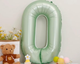 Può includere: Un grande palloncino numero zero verde chiaro. Un orsacchiotto con una felpa con cappuccio marrone è seduto accanto a un cappello di compleanno giallo con le parole "Happy Birthday". Un piccolo cestino contiene palline decorative colorate.