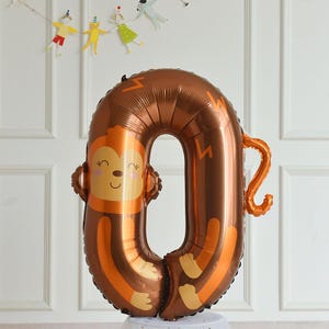 May include: A brown foil balloon shaped like the number zero with a monkey face and tail. The monkey has a brown face with orange eyes and a pink mouth.