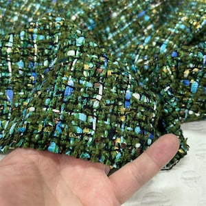 May include: Close-up of a textured fabric with a woven pattern. The fabric is primarily green with flecks of blue, white, and gold. The material appears to be thick and suitable for outerwear or upholstery.