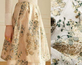 May include: A beige skirt with a floral pattern in gold and green. The skirt is made of a sheer fabric with a textured surface.