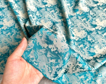 Könnte beinhalten: Nahaufnahme eines Stoffes mit türkisfarbenem und weißem Blumenmuster. Der Stoff hat einen seidigen Glanz und einen fließenden Fall. Das Design zeigt eine Mischung aus türkisfarbenen, hellblauen und weißen floralen Elementen, die einen strukturierten, künstlerischen Look erzeugen. Der Stoff ist gefaltet und zeigt die Textur und das Muster.