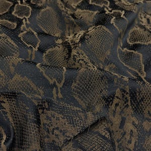 May include: A piece of fabric featuring a dark brown and black snakeskin pattern. The material has a textured surface with a repeating reptilian scale design that creates a layered visual effect across the folded and draped textile surface.