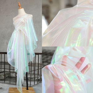 Può includere: Tessuto trasparente bianco iridescente con finitura olografica. Il tessuto è drappeggiato su un manichino e una mano tiene un pezzo di tessuto.