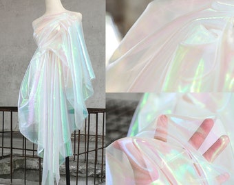 May include: White iridescent sheer fabric with a holographic finish. The fabric is draped over a mannequin and a hand is holding a piece of the fabric.
