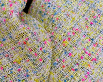 May include: Close-up of a textured fabric with a woven pattern. The fabric is primarily yellow with flecks of pink, blue, and green. The weave includes silver metallic threads, adding a subtle shimmer. The fabric appears to be a tweed or similar material.