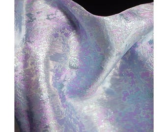 May include: Close-up of iridescent fabric with a shimmering, textured surface. The fabric features a blend of lavender, pale blue, and silver tones, creating a dynamic, flowing appearance. The material appears soft and pliable, with subtle patterns.