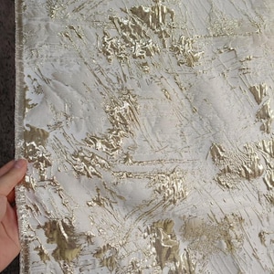 Puede incluir: Primer plano de una tela texturizada con base blanca y detalles metálicos dorados. Los elementos dorados están en relieve y crean un patrón abstracto dinámico en la superficie. La tela tiene un aspecto lujoso y decorativo.