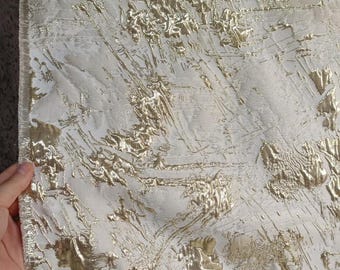 May include: Close-up of a textured fabric with a white base and gold metallic accents. The gold elements are raised and create a dynamic, abstract pattern across the surface. The fabric has a luxurious, decorative appearance.