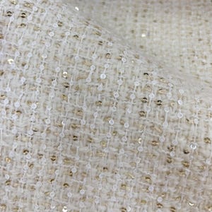 May include: A cream-colored woven fabric featuring a textured tweed-like pattern with vertical rows of small, round, white sequins interspersed with metallic gold threads throughout the material, creating a shimmering and dimensional surface appearance.