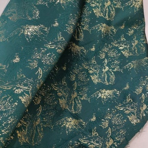 May include: Green fabric with a gold metallic print of stylized animal faces. The fabric has a textured surface.
