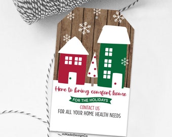 May include: A Christmas-themed gift tag with a wooden background. It features illustrations of a red house and a green house with white snow-covered roofs. The tag includes the text "Here to bring comfort home" and "FOR THE HOLIDAYS."