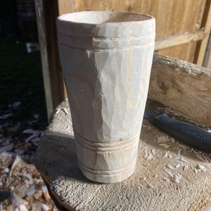 Puede incluir: Un primer plano de una taza de madera, tallada a mano a partir de una sola pieza de madera. La taza tiene una forma redonda y lisa y un acabado de madera natural. La taza est&aacute; sentada sobre una superficie de madera.