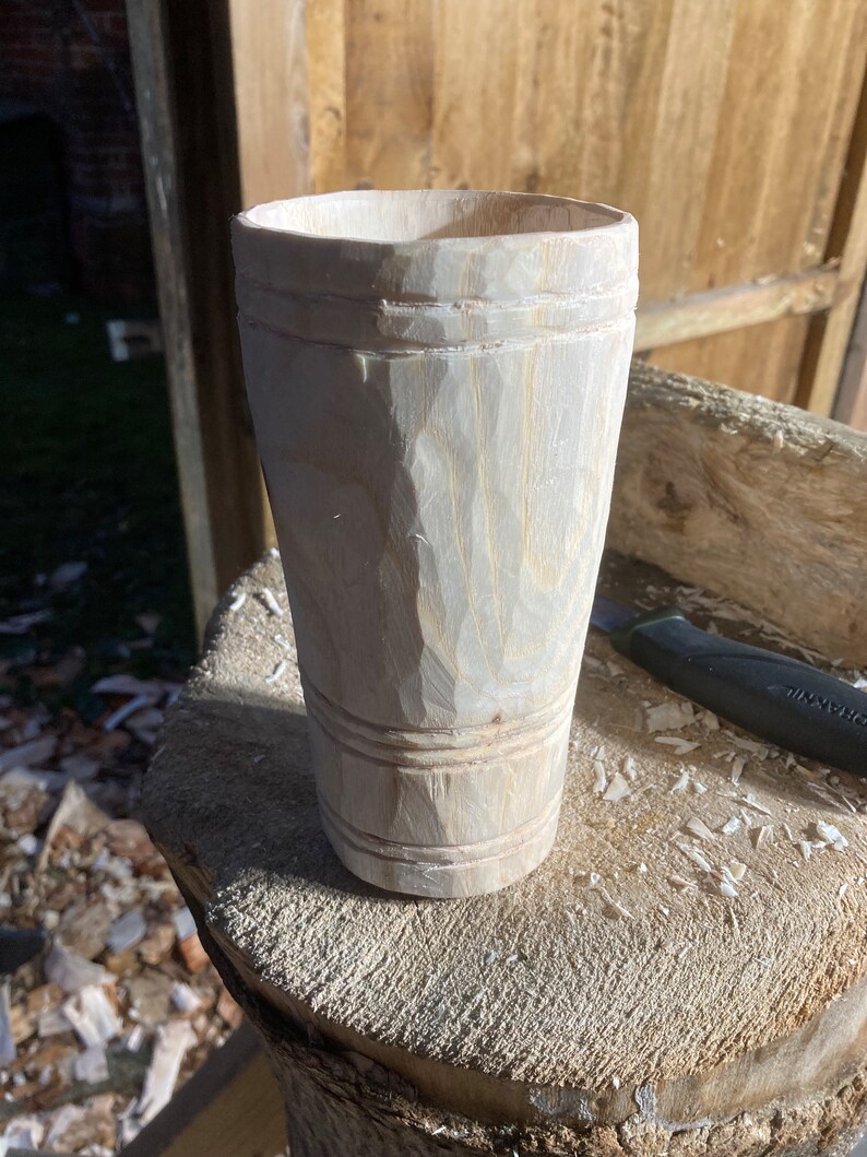 Puede incluir: Un primer plano de una taza de madera, tallada a mano a partir de una sola pieza de madera. La taza tiene una forma redonda y lisa y un acabado de madera natural. La taza est&aacute; sentada sobre una superficie de madera.