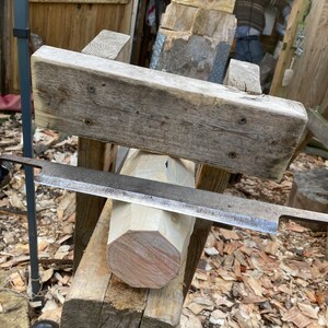 Puede incluir: Un tronco de madera se est&aacute; dando forma redonda con una herramienta de talla de madera de mano. El tronco est&aacute; asegurado en un marco de madera, y la herramienta est&aacute; apoyada en la parte superior del tronco. Se pueden ver virutas de madera en el suelo alrededor del marco.