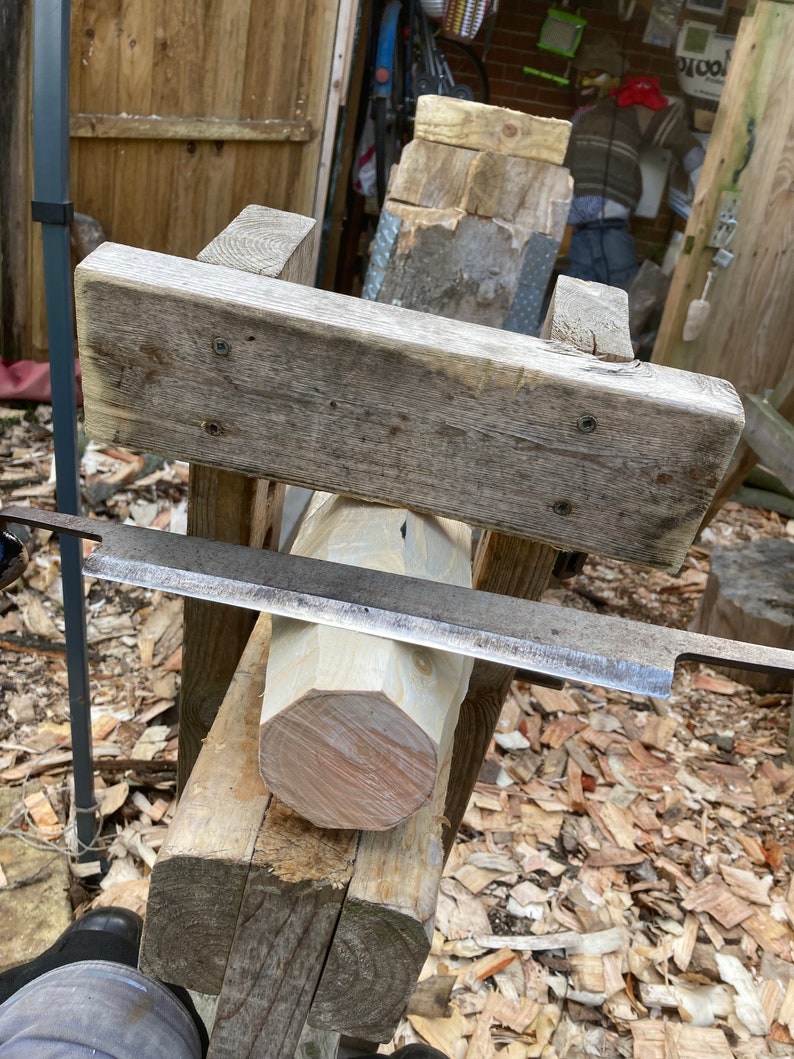 Puede incluir: Un tronco de madera se est&aacute; dando forma redonda con una herramienta de talla de madera de mano. El tronco est&aacute; asegurado en un marco de madera, y la herramienta est&aacute; apoyada en la parte superior del tronco. Se pueden ver virutas de madera en el suelo alrededor del marco.