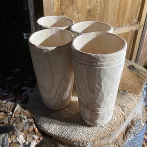 Puede incluir: Cuatro tazas de madera sin terminar, probablemente para beber, est&aacute;n dispuestas sobre un toc&oacute;n de &aacute;rbol. Las tazas son de madera clara y tienen un aspecto natural y r&uacute;stico.