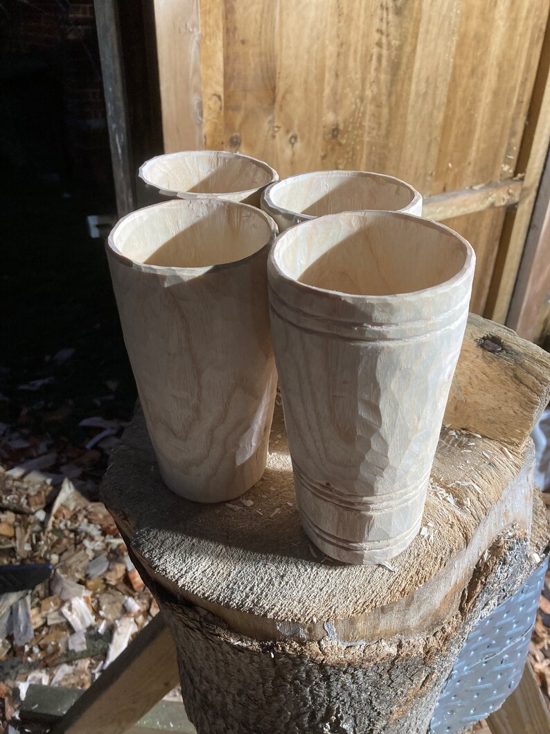 Puede incluir: Cuatro tazas de madera sin terminar, probablemente para beber, est&aacute;n dispuestas sobre un toc&oacute;n de &aacute;rbol. Las tazas son de madera clara y tienen un aspecto natural y r&uacute;stico.