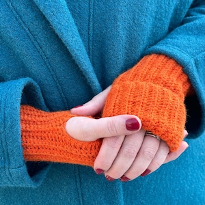 Könnte beinhalten: Ein Paar orangefarbene gestrickte Fingerhandschuhe mit Daumenloch. Die Handschuhe werden von einer Person getragen, die einen blaugrünen Mantel trägt.