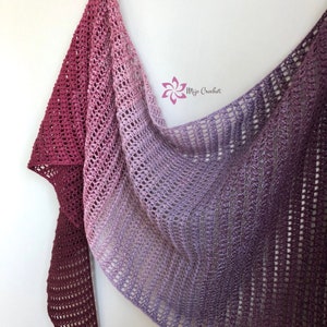May include: A crocheted shawl in a gradient of pink and purple colours. The shawl is made with a delicate open stitch pattern and has a fringed edge.