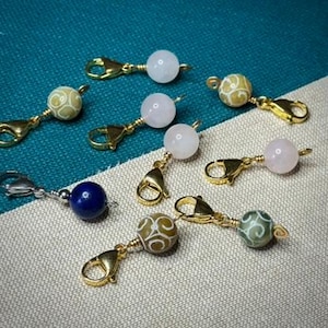 Op de afbeelding: Een verzameling bedelarmbanden met goudkleurige sluitingen. De kralen zijn in verschillende kleuren, waaronder wit, roze, blauw en groen, en sommige hebben decoratieve patronen. De bedels zijn gerangschikt op een turquoise en beige oppervlak met patroon.