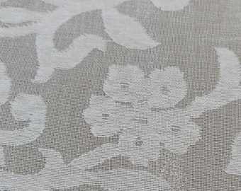 May include: Close-up of a beige fabric with a white floral pattern. The design features stylized flowers and leaves, creating a textured, elegant look. The fabric has a woven texture.