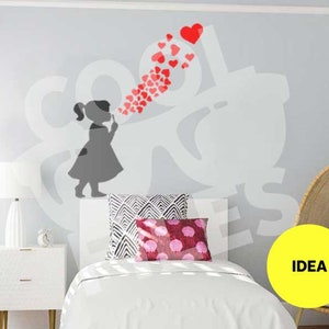 Könnte beinhalten: Ein weißes Bett mit einem weiß-schwarzen Kissen und einem rosa-weißen Kissen. Eine Silhouette eines Mädchens, das rote Herzen an eine graue Wand bläst. Der Text "IDEA" steht auf einem gelben Kreis.