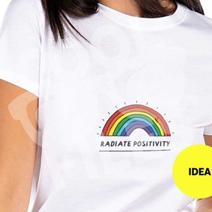 Puede incluir: Camiseta blanca con un gráfico de arcoíris y el texto "RADIATE POSITIVITY".