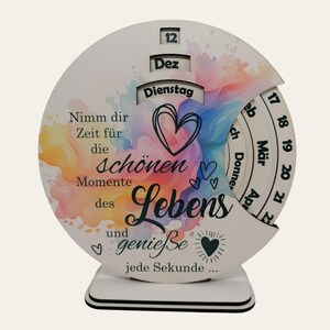 以下が含まれることがあります： 水彩画のカラフルな背景と「Nimm dir Zeit für die schönen Momente des Lebens und genieße jede Sekunde... 」というテキストが書かれた木製の手回し式カレンダー。カレンダーには、12月、2月、3月、4月の月が書かれた回転式ダイヤルがあります。