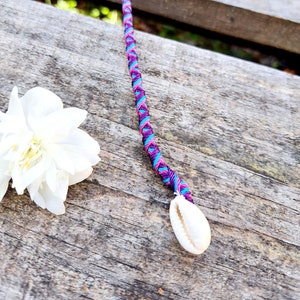 Può includere: Un cordoncino intrecciato viola, rosa e turchese con un ciondolo a forma di conchiglia bianca. Il cordoncino è appeso a una superficie di legno.